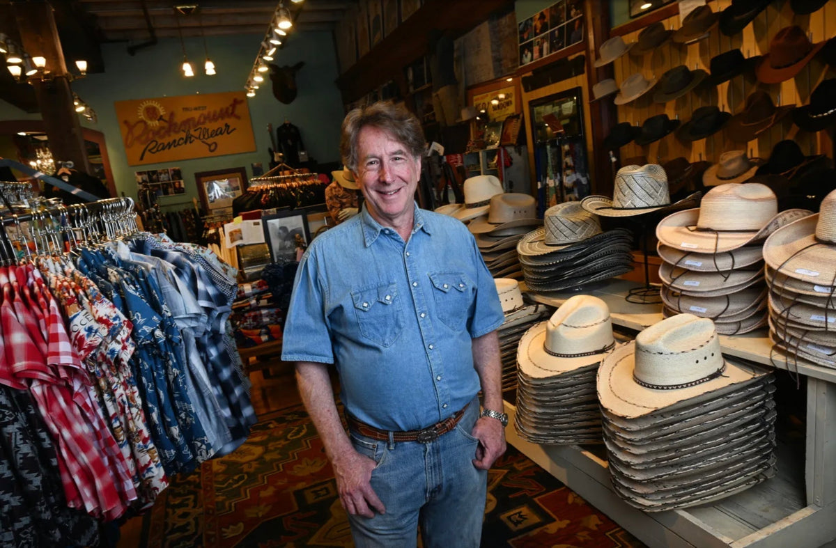 Steve Weil, president of Rockmount Ranch Wear, smiling inside the Wazee Street store surrounded by rows of Western shirts and stacked cowboy hats, wearing a classic Rockmount snap-button denim shirt.